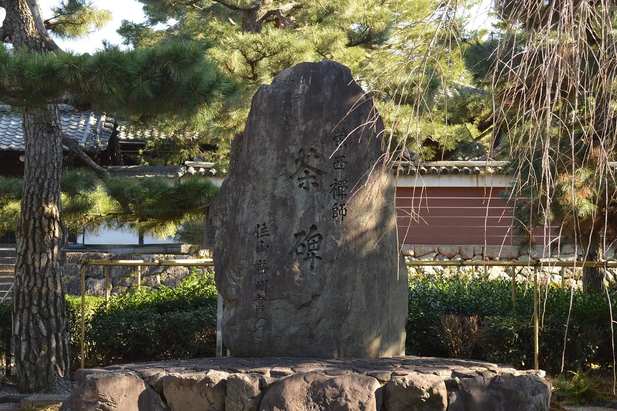 A tall carved stone monument among pine trees in front of a low red-brown wall at Kennin-ji temple in Kyoto