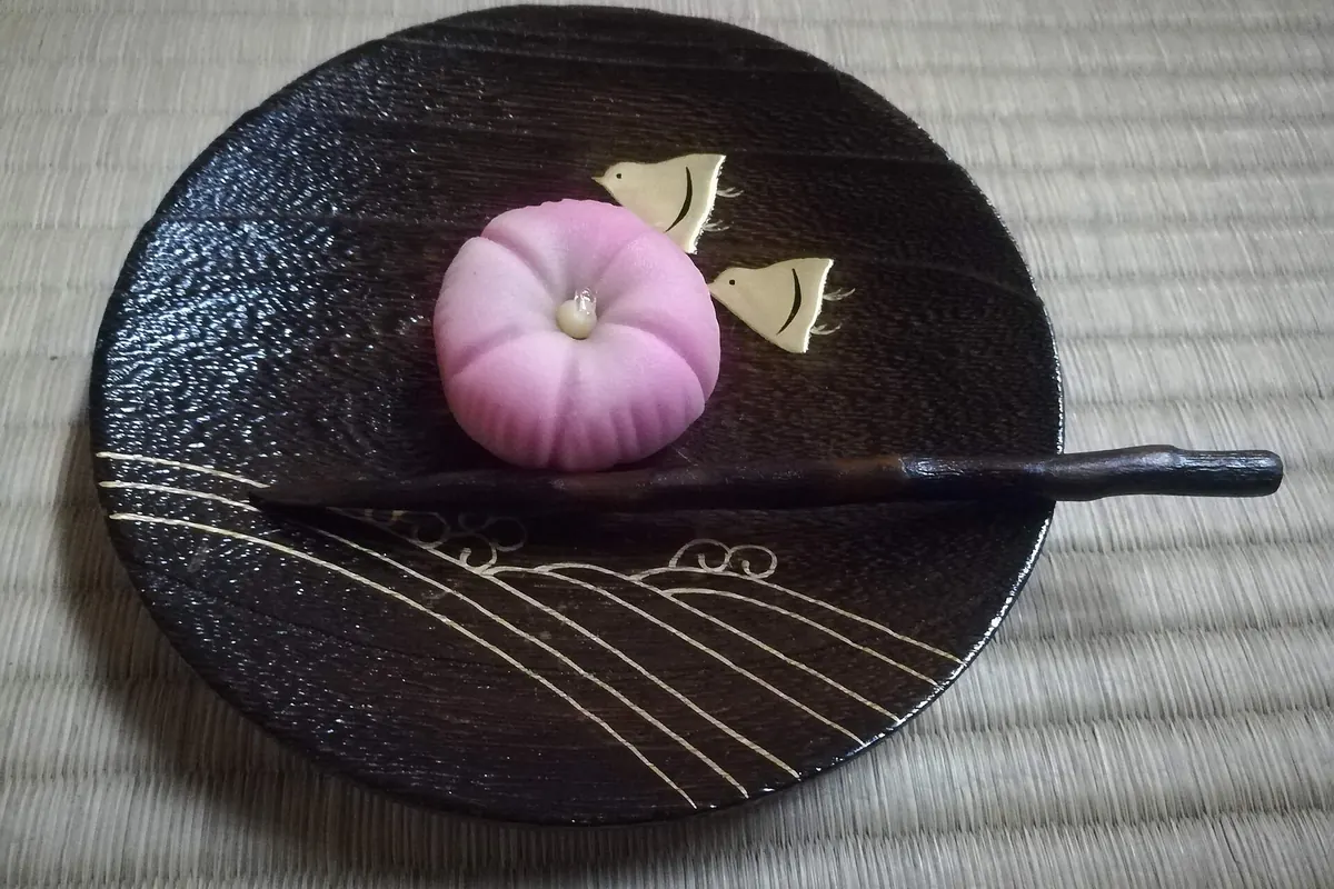 A pink flower-shaped sweet with two small metal birds on a black lacquer plate resting on a tatami mat