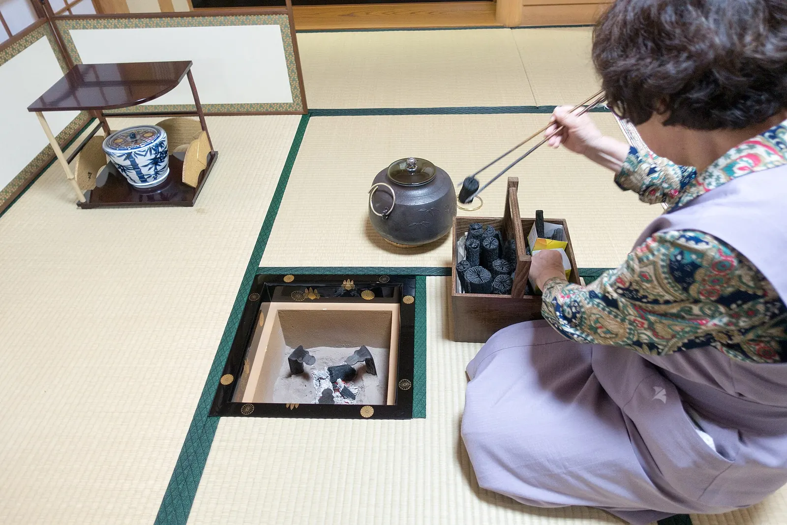 A host kneeling beside a sunken hearth, a general photograph: a woman in a lilac kimono on a tatami floor lifting charcoal with long chopsticks