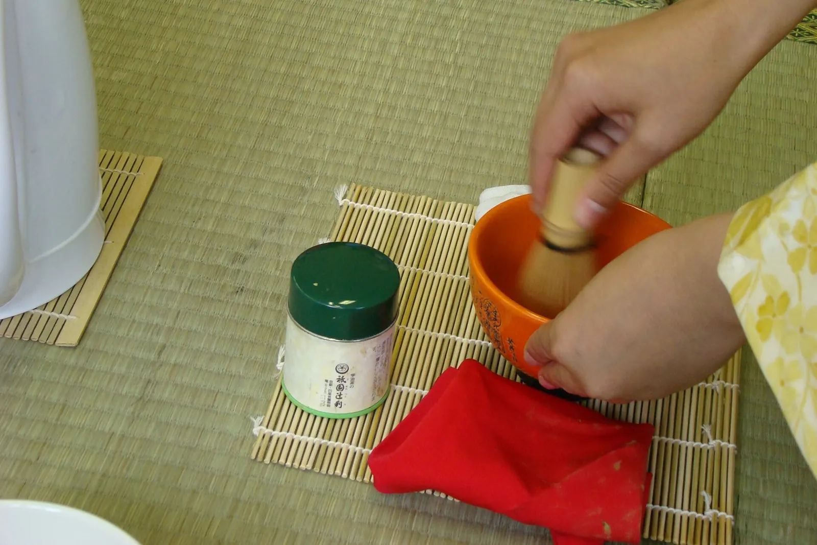 Matcha being whisked, a general photograph: a hand whisking green tea in an orange bowl on a woven mat, with a tea caddy and a red cloth beside it
